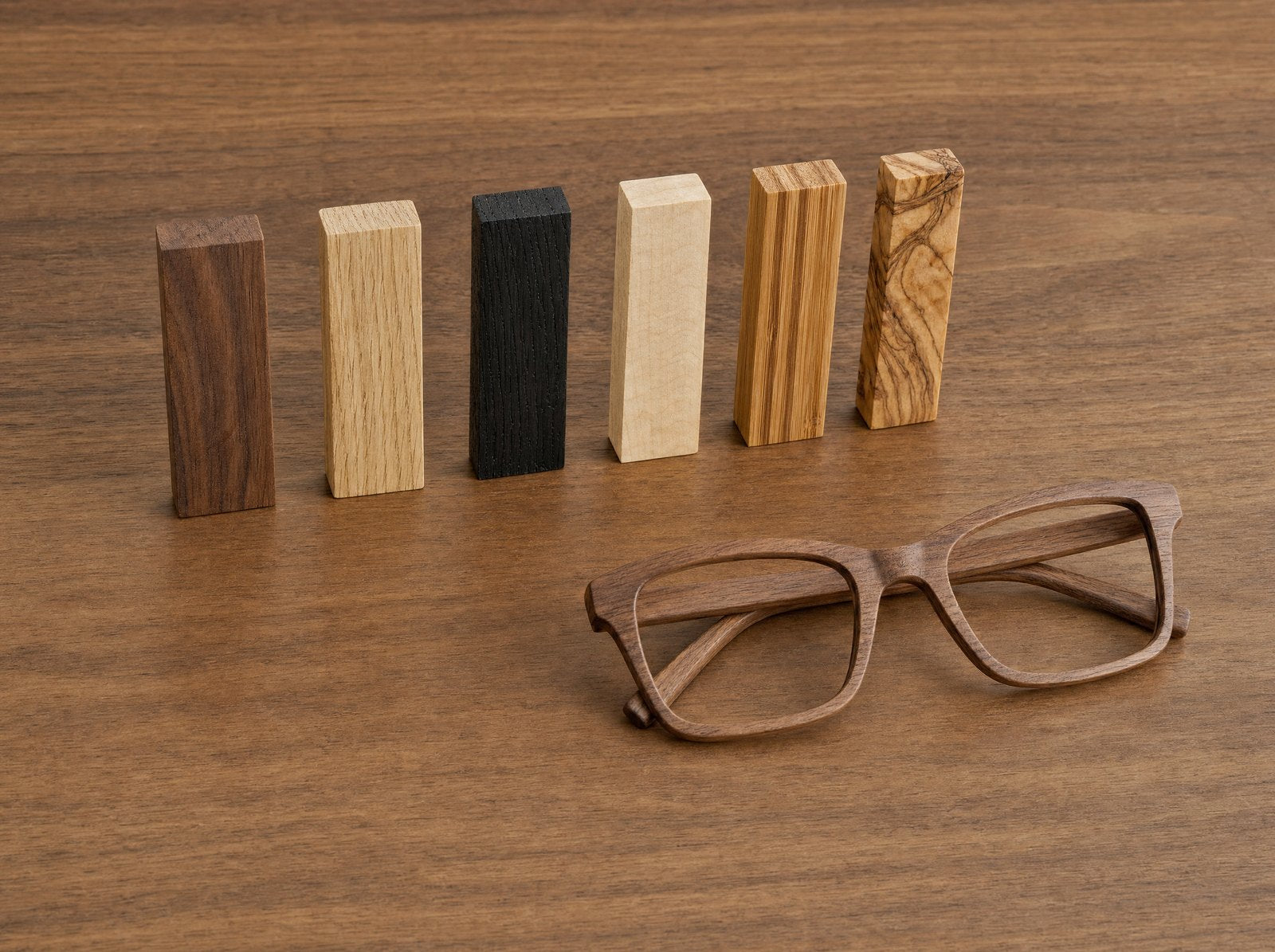 Sechs Holzmuster verschiedener Arten nebeneinander, daneben eine fertige Holzbrille